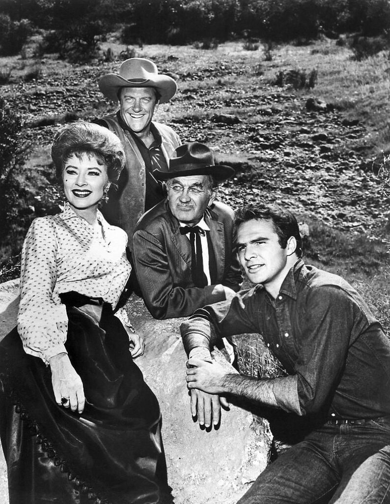 Photo of the main cast for the television program Gunsmoke in 1963. From left: Amanda Blake (Miss Kitty), James Arness (Matt Dillon), Milburn Stone (Doc Adams), and Burt Reynolds (Quint Asper).